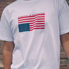 White t-shirt with American flag graphic upside down and 'AMERICA IS REVOLTING' text on a blurred brick wall background