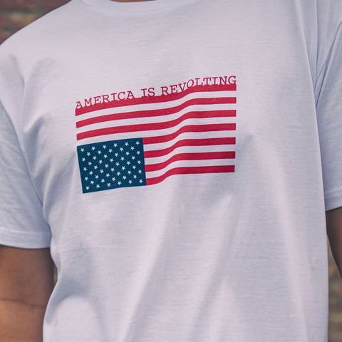 White t-shirt with American flag graphic upside down and 'AMERICA IS REVOLTING' text on a blurred brick wall background