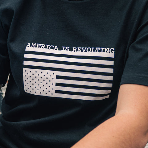 Black t-shirt with white American fkag graphic upside down and text 'AMERICA IS REVOLTING' on a person.