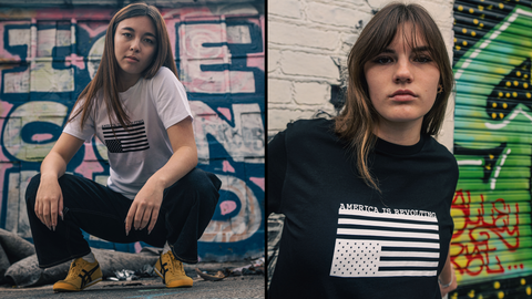 Woman in white t-shirt with a black print and woman in black t-shirt with a white print where both shirts have an American flag graphic upside down and 'AMERICA IS REVOLTING' text 